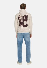 Sudadera crema con un estampado gráfico en la parte posterior que presenta diseños abstractos. Combinada con jeans de denim azul claro y zapatillas blancas.