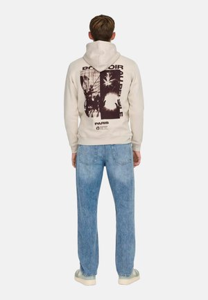 Sudadera crema con un estampado gráfico en la parte posterior que presenta diseños abstractos. Combinada con jeans de denim azul claro y zapatillas blancas.