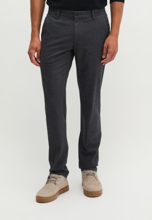 Mann trägt dunkelgraue karierte Hose und hellbeige Wildleder-Schnürschuhe und steht vor einem schlichten Hintergrund.