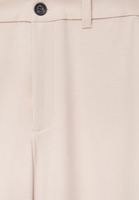 Pantalon beige avec une texture de tissu lisse, présentant une fermeture à bouton et une poche latérale. Les détails cousus sont subtils et épurés.