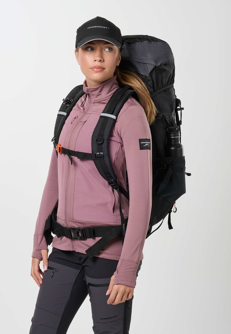 Femme portant une veste mauve, un pantalon de randonnée noir, une casquette noire et un grand sac à dos noir avec une bouteille d'eau, regardant vers la droite sur un fond blanc.