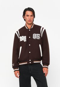 Giacca varsity marrone con strisce bianche e dettagli ricamati, dotata di chiusura a bottoni a pressione e polsini a costine. Il testo sulla manica recita "Fondata nel 1985".