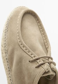 Beige mockasin i mocka med synliga sömmar och snören, med en vävd sula mot en neutral bakgrund.