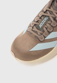 Καφέ αθλητικό παπούτσι με τόνους ανοιχτού μπλε, υλικό πάνω μέρος με υφή και επίπεδη μπεζ σόλα. Διαθέτει κορδόνια και σήμανση στο πλάι.