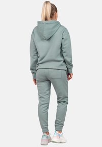 Felpa verde chiaro e joggers coordinati in tessuto morbido. L'outfit presenta polsini a coste e una tasca posteriore sui pantaloni. Le sneaker sono multicolori.