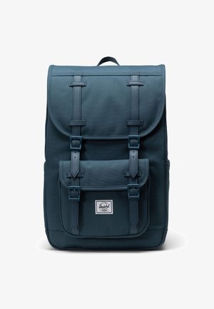 Mochila azul oscura con tapa de solapa, dos hebillas en el frente y un bolsillo frontal. Hecha de un tejido resistente, con un parche de logo en la parte inferior.