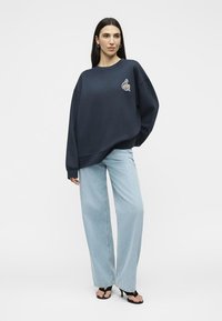 Femme portant un sweat-shirt ample bleu marine avec un petit motif sur la poitrine, un jean large bleu clair, et des sandales noires à talons.