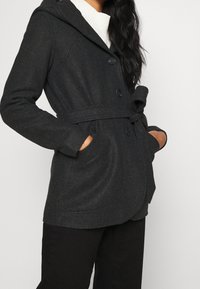 Abrigo peacoat gris oscuro con capucha, cierre de botones, cintura ceñida con cinturón, bolsillos laterales y una textura suave. Combinado con un suéter blanco debajo.