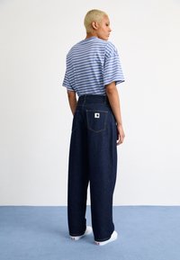 Pantalons en denim bleu foncé, taille haute et ample, avec des poches arrière. Associés à une chemise à manches courtes à rayures bleues et blanches.