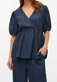 Blouse bleu foncé avec manches courtes bouffantes, encolure en V et silhouette péplum. Tissu texturé avec des rayures verticales.