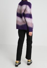 Pull en peluche rayé violet et rose, coupe décontractée, assorti à un pantalon noir et des chaussures grises avec des accents jaunes. Texture douce, look superposé.