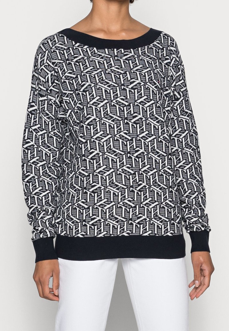 Pull à manches longues noir et blanc avec un motif géométrique, porté par une personne avec un pantalon blanc sur un fond uni.