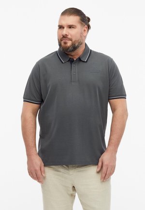 Man with beard and tied hair wearing a dark gray polo shirt and light beige pants, looking sideways against a plain background.