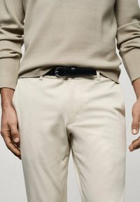 Pull en tricot beige avec manches longues associé à un pantalon crème et une ceinture en cuir noir. Vue rapprochée mettant l'accent sur la taille.
