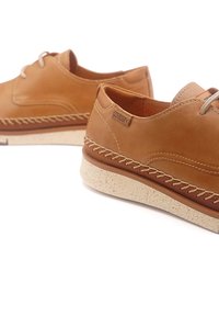 Des chaussures en cuir marron avec des détails de couture, des lacets ronds et une semelle en caoutchouc crème texturée. Logo de la marque visible sur le côté.