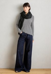 Stribet langærmet bluse i sort og hvid, parret med brede mørke denimjeans. Modellen bærer et chunky sort tørklæde og står på et trægulv.