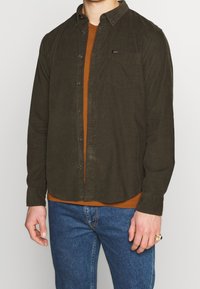 Mörkgrön skjorta med knappar, krage, bröstficka och texturerat tyg över en brun skjorta. Kombinerad med blå jeans och en ring.