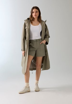 Femme portant un long manteau vert olive sur un short ceinturé assorti et un débardeur blanc, debout devant un fond clair uni.