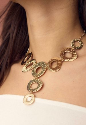 Primo piano di una persona che indossa una collana in oro martellato con cerchi aperti irregolari collegati su pelle nuda e top bianco.