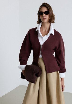 Frau mit schwarzen Sonnenbrillen, weißem Hemd mit Kragen, bordeauxrotem Cardigan, beigem Rock, hält eine gefaltete bordeauxrote Jacke vor einfarbigem Hintergrund.