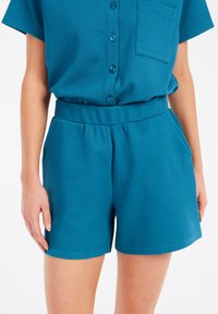 Mėlynos šortai iš tekstūruoto audinio su elastingu juosmeniu, turintys šonines kišenes ir laisvą siluetą. Viršuje yra detalės su mygtukais.