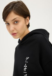 Noor inimene lühikeste pruunide juustega kannab musta kapuutsiga pusa, millel on valged vertikaalsed jaapani tekstid, taust on ühevärviline hele.