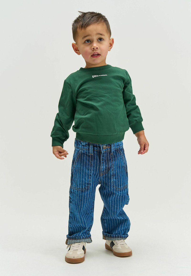 Jeune garçon debout, portant un sweat-shirt vert avec le texte « DREAMER », un jean rayé bleu et des baskets blanches sur un fond clair uni.