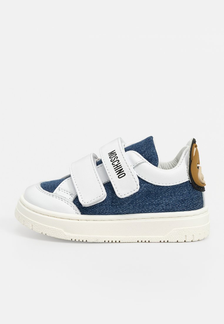 Baskets en denim avec des accents synthétiques blancs, deux sangles Velcro et un détail d'ours brun sur le talon. Tissu bleu texturé et semelle en caoutchouc.