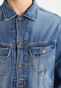 Džínová bunda světle modré barvy s špičatým límcem, zapínáním na knoflíky a dvěma předními kapsami. Výrazné šití a kovové knoflíky.