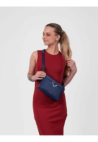 Námornícká crossbody taška s elegantním obdélníkovým tvarem, matným povrchem, V-čkovým kovovým akcentem a nastavitelným popruhem, nošena přes červené šaty.