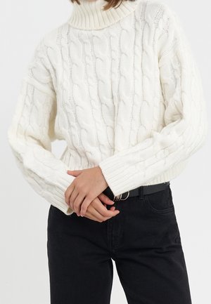 Frau trägt einen weißen, grob gestrickten Rollkragenpullover mit langen Ärmeln und schwarze, hoch taillierte Hosen, die Hände vor der Taille verschränkt.