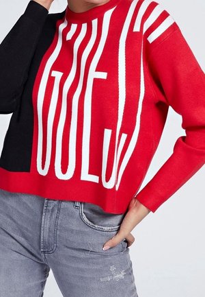 Osoba ubrana w sweter w kolorach czerwonym, czarnym i białym z dużymi pionowymi literami oraz jasnoszare dżinsy z subtelnym efektem przetarcia.