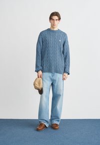 Μπλε πουλόβερ με πλέξη καλωδίου και μανσέτες με ρίγες, ανοιχτό μπλε τζιν, καφέ παπούτσια και μπεζ καπέλο κρατημένο στο χέρι σε απλό φόντο.