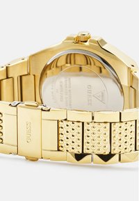 Reloj de acero inoxidable dorado con una correa texturizada que presenta patrones hexagonales y una parte trasera plateada, que incluye una corona para ajustar la hora.