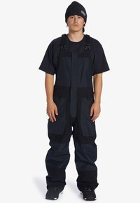 Zwarte geïsoleerde sneeuwoverall met verstelbare banden, meerdere zakken, een ontspannen pasvorm en duurzaam materiaal. Draagt over een zwart t-shirt.