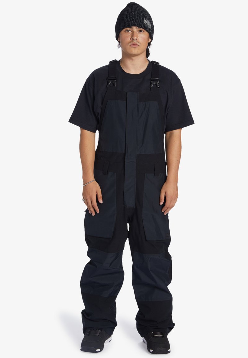 Zwarte geïsoleerde sneeuwoverall met verstelbare banden, meerdere zakken, een ontspannen pasvorm en duurzaam materiaal. Draagt over een zwart t-shirt.