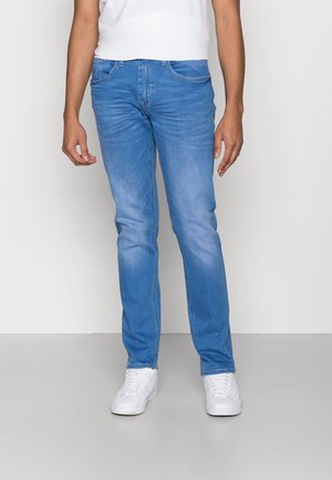 Homme portant un jean bleu clair slim-fit et des baskets blanches, debout devant un fond gris clair uni.