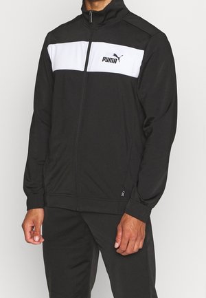 Mężczyzna ubrany w czarną kurtkę sportową Puma na zamek z białym poziomym paskiem na klatce piersiowej oraz czarne spodnie.