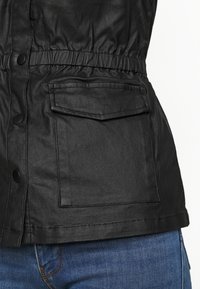 Musta tekonahkainen takki, jossa on joustava vyötärö, nepparikiinnitys ja edessä sileäpintainen tasku. Yhdistetty sinisiin farkkuihin.