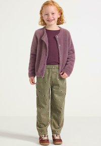 Cardigan viola peloso sopra una maglietta viola scura, pantaloni verdi con motivo floreale e scarpe da ginnastica marroni.