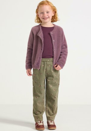 Cardigan viola peloso sopra una maglietta viola scura, pantaloni verdi con motivo floreale e scarpe da ginnastica marroni.