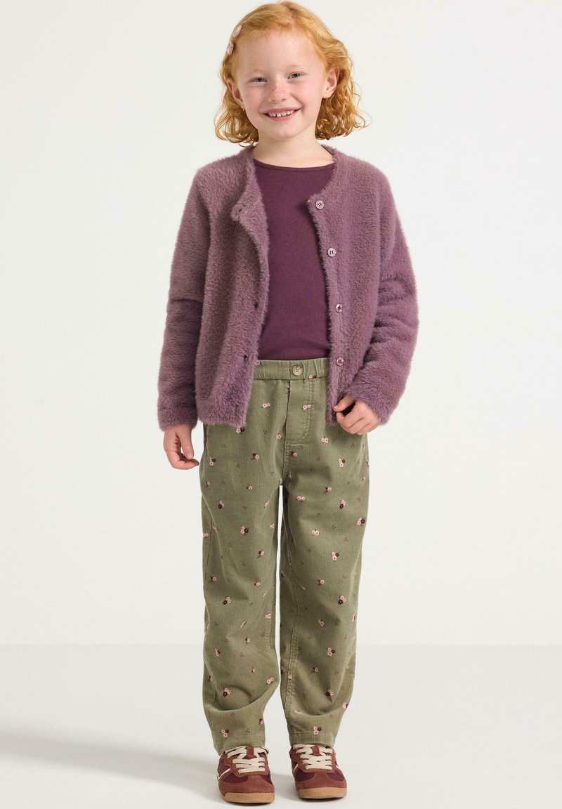Cardigan viola peloso sopra una maglietta viola scura, pantaloni verdi con motivo floreale e scarpe da ginnastica marroni.