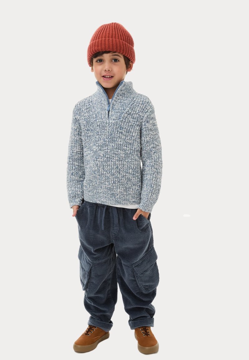 Maglione lavorato a maglia azzurro chiaro con colletto a mezza zip, abbinato a pantaloni cargo in velluto a coste blu scuro e scarpe stringate marroni. Beanie lavorato a maglia rosso.