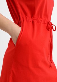 Robe en coton rouge avec une taille cintrée, dotée de poches latérales et de détails à cordon ajustable à la taille. Texture lisse.
