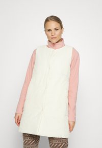Colete acolchoado branco com gola alta, apresentando botões de pressão e bolsos laterais, usado sobre uma blusa de manga longa rosa e leggings estampadas.