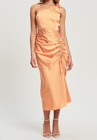 Robe en satin pêche à épaule dénudée avec des détails froncés et un lien sur le côté. Longueur mi-mollet avec une texture lisse et une silhouette ajustée. Talons transparents.