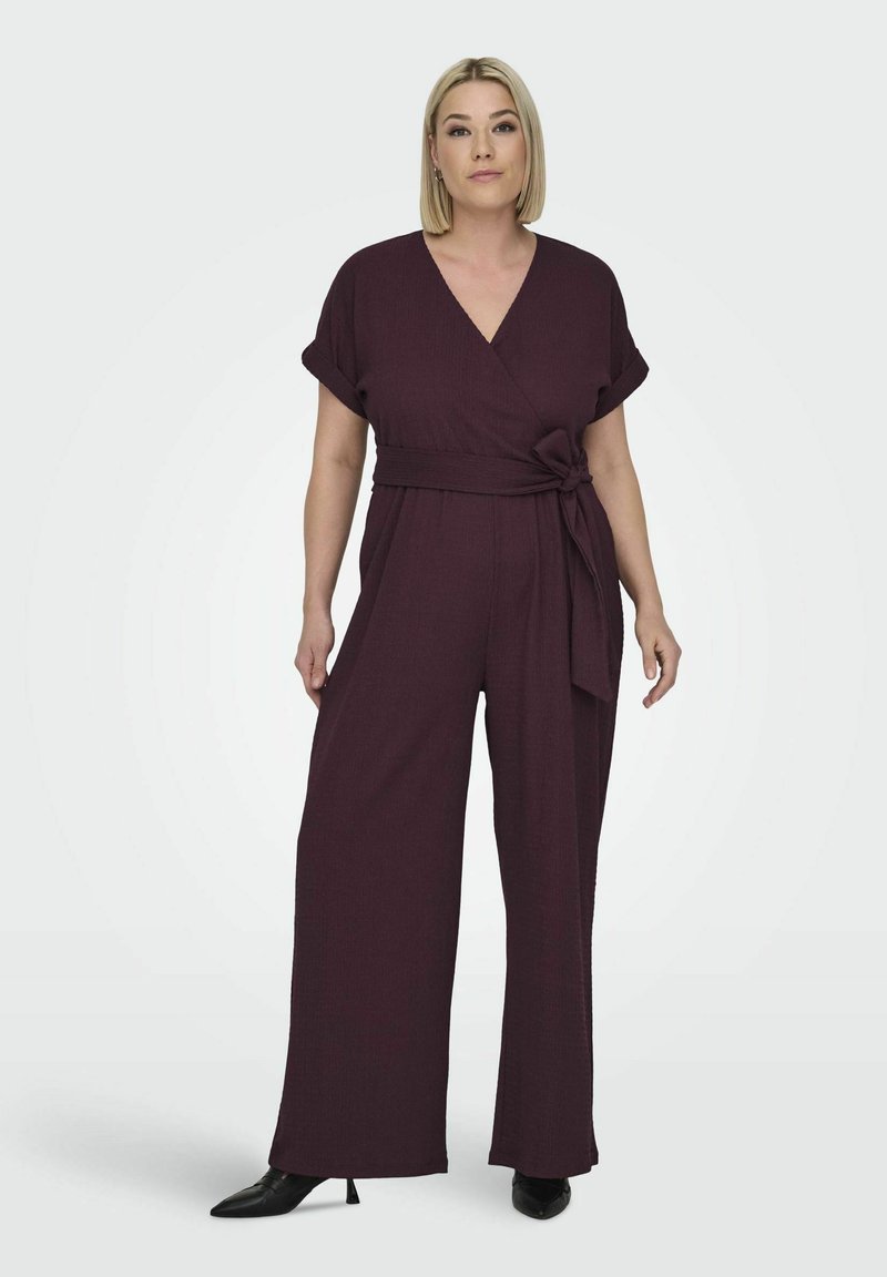 Bordový zavinovací overal s krátkymi rukávmi, textúrovaná látka, široké nohavice a viazanie v páse na úpravu. Čierne špicaté topánky.