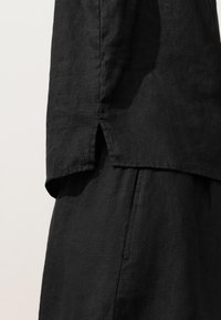 Gros plan sur un haut en lin noir et un pantalon montrant un détail de fente latérale sur l’ourlet du haut sur un fond neutre.