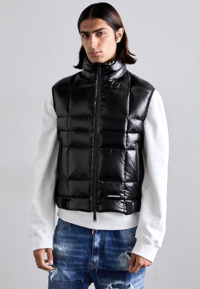 Sort puffer vest med kvadratisk quiltet design, høj krave, lynlås foran og logo detalje. Båret over en hvid langærmet top.