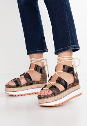 Sandales plates-formes avec dessus en tissu camouflage marron et noir, semelle intermédiaire en jute tressé et semelle extérieure en caoutchouc blanc. Fermeture à lacets réglable.